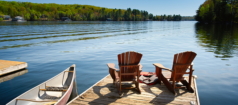 Two Adirondack chairs on a wooden dock facing the blue water of a lake in Muskoka, Ontario Canada. Life jackets are visible near the chairs. A canoe is tied to the pier, paddles are stored inside.